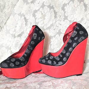 May include: A pair of black wedge heels with a skull pattern and bright red platforms. The shoes have a satin ribbon detail and a high wedge heel. The shoes are displayed on a white floral patterned fabric.