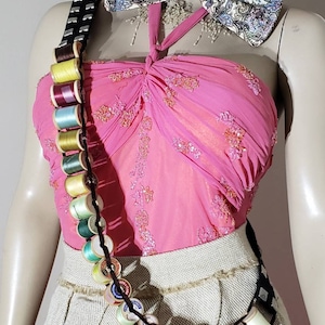 May include: A pink halter top with floral embroidery, a beige skirt, and a bandolier-style strap adorned with various colorful sewing thread spools. A large paisley-patterned bow tie is worn at the neck, and a pink hat is visible at the top.