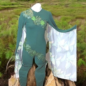 May include: A green jumpsuit with long sleeves and attached cape. The jumpsuit features leaf-like embellishments on the chest and leg. The cape is white with a green leaf pattern. The outfit is displayed on a wooden surface against a backdrop of greenery.