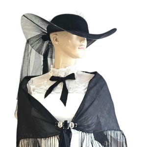 May include: A black hat with a wide brim and a sheer veil, paired with a black shawl featuring fringe and a decorative clasp. The ensemble is displayed on a mannequin, showcasing a vintage or gothic aesthetic.