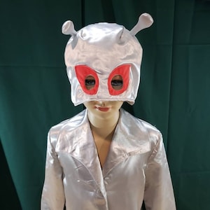 May include: A shiny silver jacket paired with a matching alien-themed headpiece. The headpiece features two protruding antennae and vibrant red eye-shaped cutouts. The jacket has a classic lapel collar and a button-down front closure.
