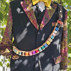 May include: A black blazer with a vibrant purple and yellow paisley pattern on the lapels and sleeves, paired with a yellow bow tie. A bandolier made of colorful thread spools is worn across the chest, and a silver chain hangs from the front button.