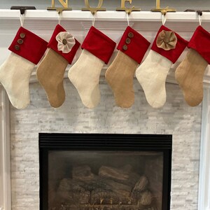 May include: Six burlap Christmas stockings with red felt cuffs and various embellishments, including buttons, a flower, and a bow, hanging on a white mantel above a fireplace.