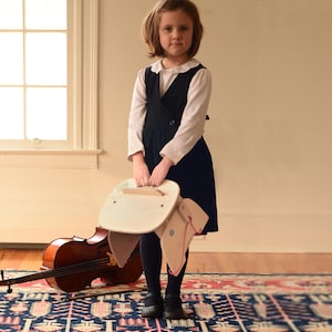 May include: A light-colored wooden stool with a curved seat and a red string detail, held by a child wearing a white long-sleeved shirt, a dark navy blue pinafore dress, and dark tights, with a brown wooden cello resting on the floor nearby.