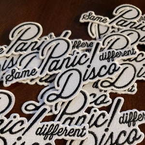 May include: A close-up of several white embroidered patches with black lettering. The patches read "Panic! Disco" and "Same Different".