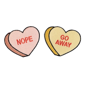 May include: Two heart-shaped candy conversation hearts with embroidered messages. The pink heart says "NOPE" and the yellow heart says "GO AWAY".