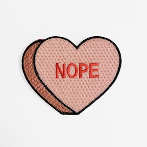Puede incluir: Un caramelo en forma de corazón rosa con la palabra "NOPE" bordada en rojo.