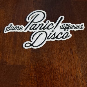 May include: White embroidered patch with black lettering that reads "Panic! Disco" with the words "same" and "different" on either side of the text.