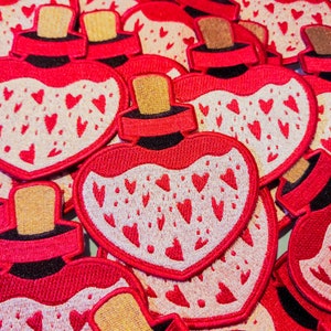 May include: A close-up of several red heart-shaped fabric patches with white embroidered hearts and a gold top. The patches are sewn together to create a layered effect.