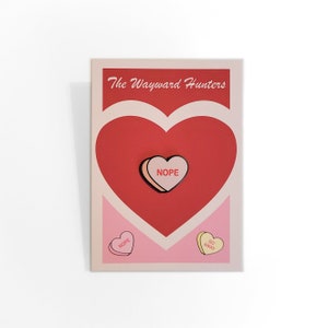 Puede incluir: Un pin en forma de corazón rosa y blanco con la palabra "NOPE" en letras rojas. El pin está en una tarjeta con un corazón rojo y el texto "The Wayward Hunters". Hay otros dos pines en la tarjeta, uno rosa con la palabra "NOPE" y uno amarillo con el texto "GO AWAY".