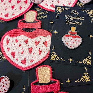 Puede incluir: Una botella roja en forma de corazón llena de corazones rosas y una tapa dorada. La botella es un parche con detalles bordados. El texto "The Wayward Hunters" está sobre un fondo negro con estrellas doradas y un borde dorado.
