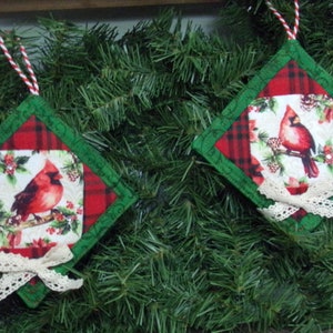 May include: Two red cardinal Christmas ornaments with plaid and green fabric, white lace trim, and a red and white twine hanger.