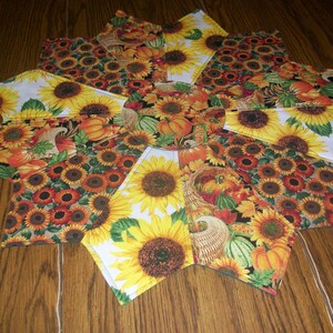 May include: A round table runner with a sunflower and autumn harvest design. The runner is made of fabric and has a white background with yellow, orange, and brown accents.