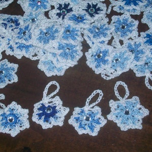 May include: A collection of crocheted snowflake ornaments in white and blue with sparkly accents. The snowflakes are arranged on a brown surface.