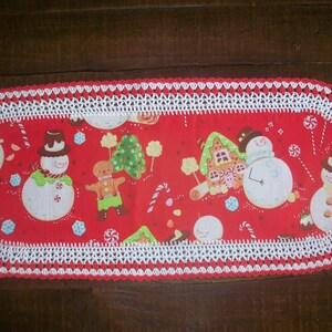 May include: A red oval table runner with a white crocheted border. The runner features a festive Christmas design with snowmen, gingerbread people, candy canes, and a gingerbread house.