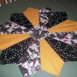 May include: A round table runner with a black, white, and yellow rooster print. The runner is made of fabric and has a starburst design.