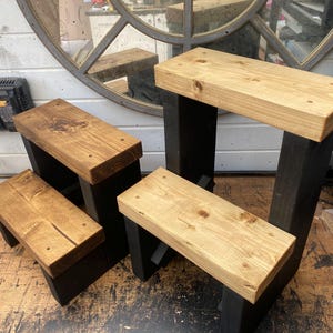 May include: Four wooden step stools with natural wood tops and black legs are displayed. The stools vary in height and size, showcasing the natural wood grain. The background includes a mirror and a tool case.