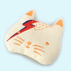May include: White fabric cat head pillow with orange ears and embroidered details. The cat has a red and blue lightning bolt design on its forehead and orange whiskers.