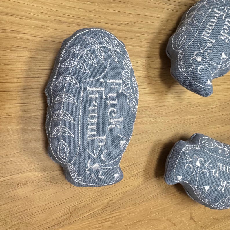 May include: Three blue fabric ornaments with white embroidery. The ornaments are shaped like a teacup and have the text "Tuck in,  Truffle" embroidered on them.