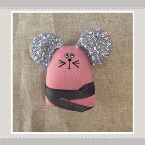 May include: A pink fabric mouse with black embroidered eyes and whiskers. The mouse has large ears covered in a white and gray floral fabric.