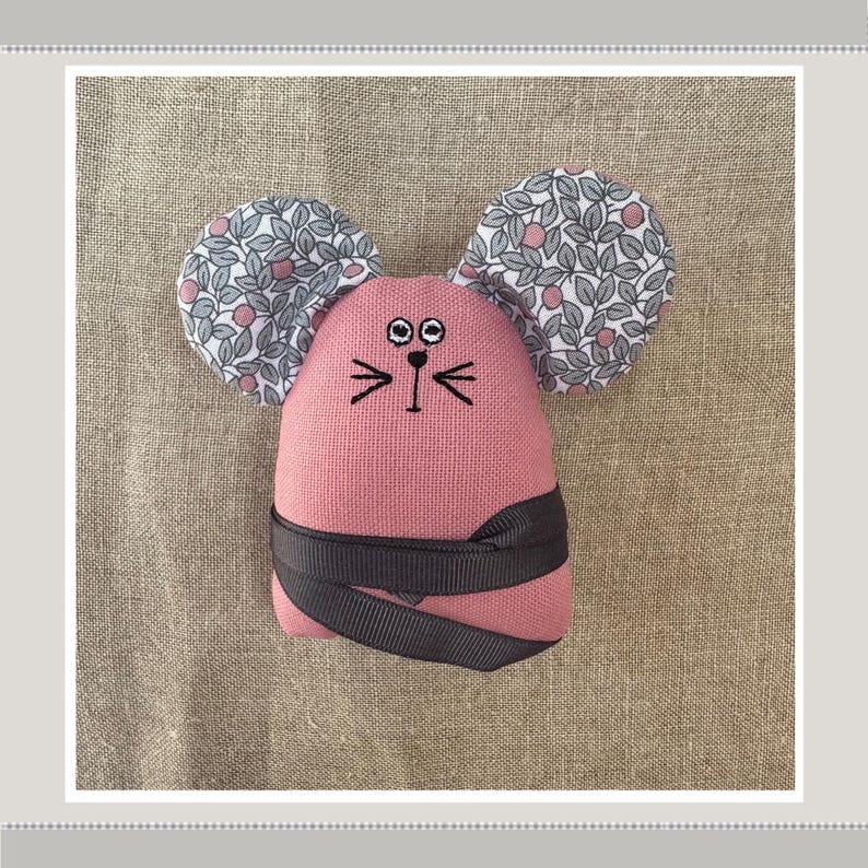 May include: A pink fabric mouse with black embroidered eyes and whiskers. The mouse has large ears covered in a white and gray floral fabric.