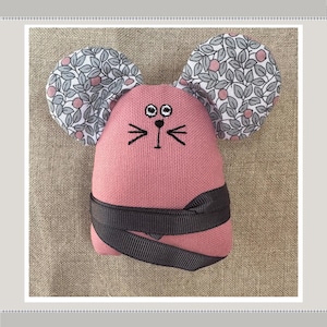 May include: A pink fabric mouse with black embroidered eyes and whiskers. The mouse has large ears covered in a white and gray floral fabric.