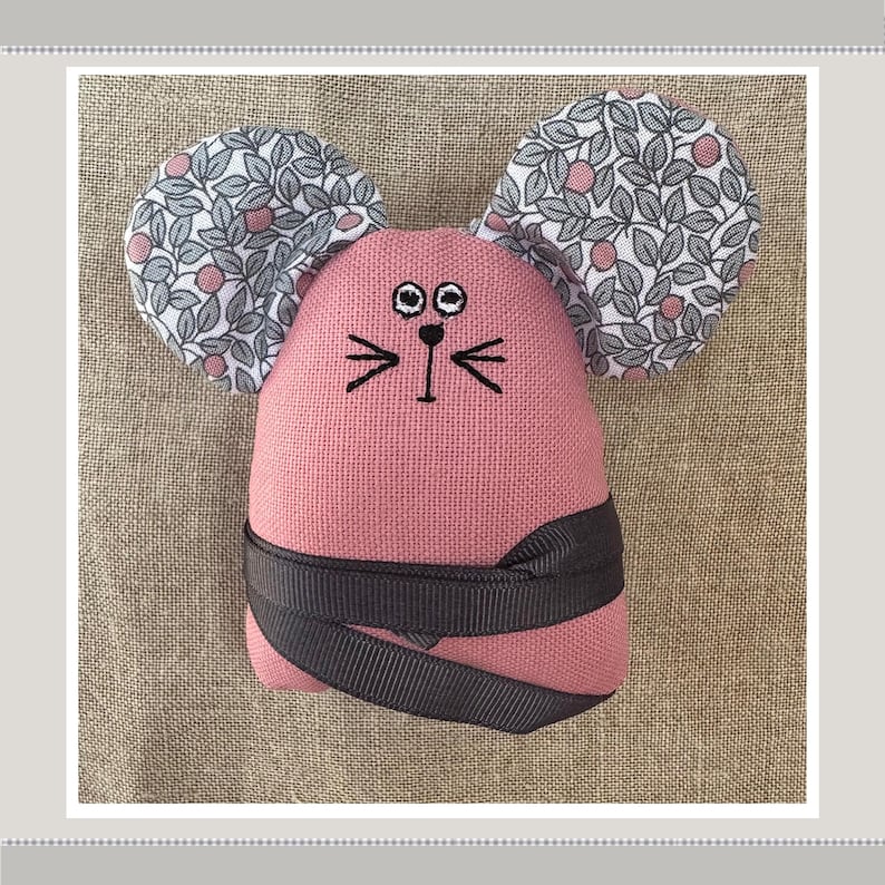 May include: A pink fabric mouse with black embroidered eyes and whiskers. The mouse has large ears covered in a white and gray floral fabric.