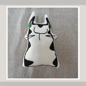 May include: A white fabric cat toy with black and green embroidered details. The cat has a simple, cartoon-like design with large eyes and a smiling mouth.