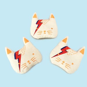 May include: Three white plush cat-shaped toys with embroidered details. Each toy features a red and blue lightning bolt design and a yellow embroidered face.