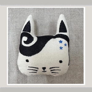 May include: A white plush toy shaped like a cat with black embroidered features, including a swirl on one side and blue stars on the other.