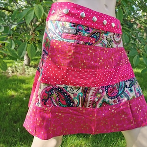 May include: A pink and white patchwork skirt with a paisley print and gold accents. The skirt has a waistband with silver buttons and a small pocket with the brand name "Mori" printed on it.