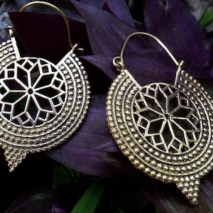 Könnte beinhalten: Ein Paar goldfarbene Ohrringe. Jeder Ohrring hat ein kreisförmiges Design mit einem sternförmigen Ausschnitt in der Mitte, umgeben von einem gepunkteten Rand. Die Ohrringe haben einen Hakenverschluss und einen dekorativen, spitz zulaufenden Boden.