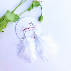 May include: White feather earrings with crystal accents. The earrings are handmade and feature a delicate feather design. The earrings are displayed on a white background with a pink flower.