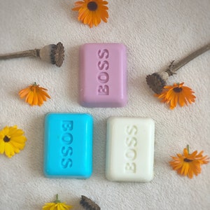 Op de afbeelding: Drie stukken zeep met het woord "BOSS" erop gedrukt. De zeep is roze, blauw en wit.