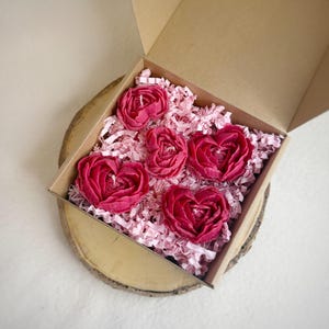 以下が含まれることがあります： 6つのハート型の濃いピンク色のキャンドルが入った段ボール箱。キャンドルはピンク色の紙吹雪の中にあります。箱は木の輪切りに置かれ、背景はニュートラルです。