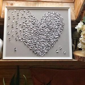 May include: A white framed wall art piece featuring a heart shape made of gray paper butterflies.