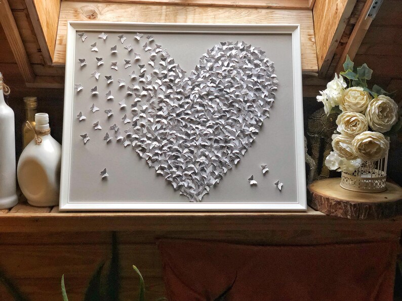 May include: A white framed wall art piece featuring a heart shape made of gray paper butterflies.