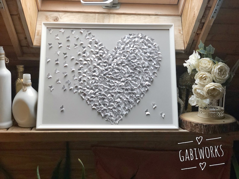 May include: A white framed canvas with a heart shape made of gray paper butterflies. The heart is on a gray background. The canvas is hanging on a wall with a wooden surface.