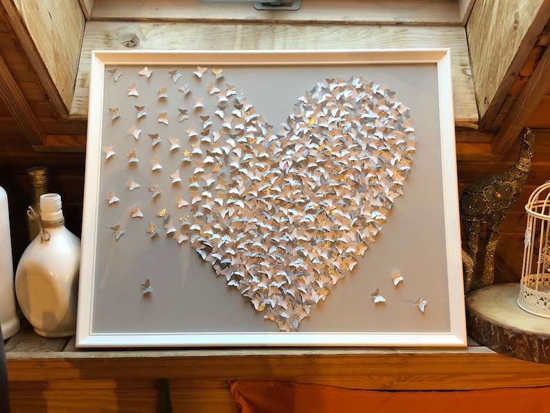 May include: A white frame with a gray background featuring a heart shape made of silver butterflies. The butterflies are arranged in a way that creates a 3D effect.