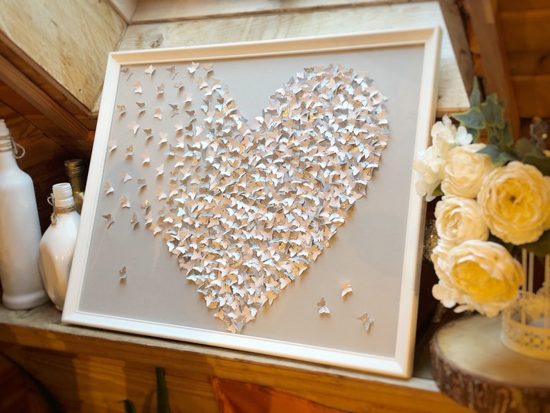 May include: A white frame with a gray background and a heart shape made of silver and white butterflies. The heart is centered in the frame.