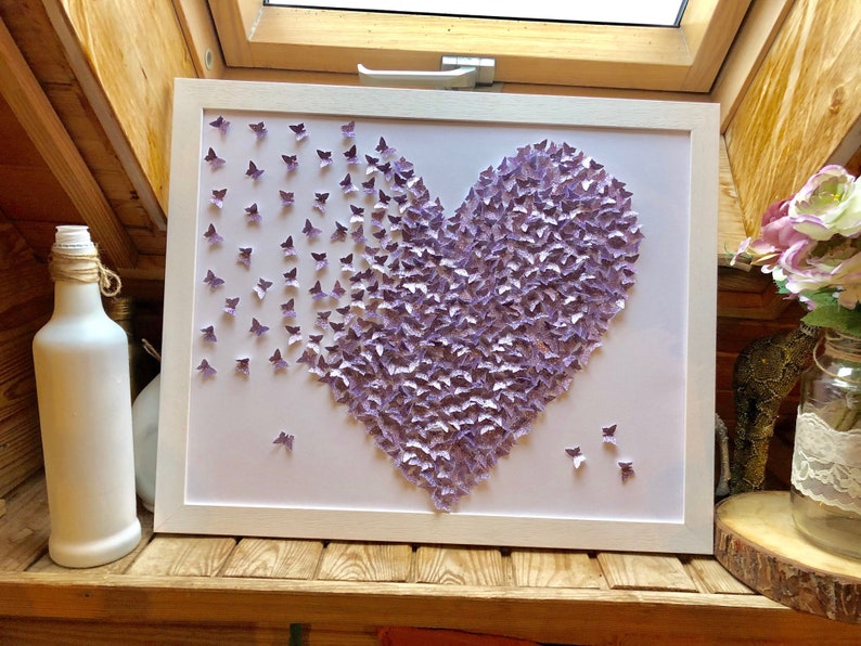 Op de afbeelding: Een wit ingelijst canvas met een hartvorm gemaakt van paarse vlinders. De vlinders zijn gerangschikt in een verloop, met de donkerste vlinders in het midden van het hart en de lichtste vlinders aan de randen.