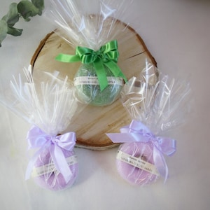 Könnte beinhalten: Drei runde Badebomben, verpackt in durchsichtigem Cellophan mit einem Band um jede. Die Badebomben sind grün, lila und lavendelfarben. Die Etiketten auf den Badebomben lauten "Vannas Zumbo", "Ziedu Skola" und "Vasara C Ziedu Garša".