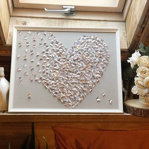 May include: A framed heart-shaped wall art piece made of silver and gold butterflies. The butterflies are arranged in a heart shape on a light gray background.