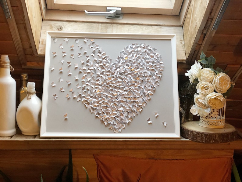 May include: A framed heart-shaped wall art piece made of silver and gold butterflies. The butterflies are arranged in a heart shape on a light gray background.