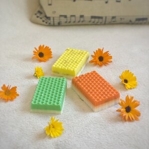 Può includere: Tre saponette con un motivo a nido d'ape. Le saponette sono verde, giallo e arancio. Le saponette sono circondate da piccoli fiori arancioni.