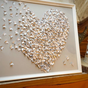 May include: A white frame with a gray background featuring a heart shape made of silver and white butterflies. The butterflies are arranged in a gradient pattern, with the silver butterflies clustered in the center and the white butterflies scattered around the edges.