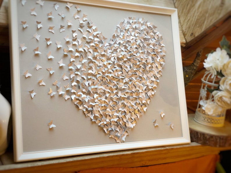 May include: A white frame with a gray background featuring a heart shape made of silver and white butterflies. The butterflies are arranged in a gradient pattern, with the silver butterflies clustered in the center and the white butterflies scattered around the edges.