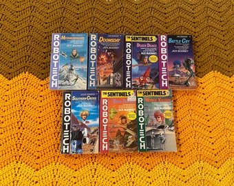 May include: A collection of eight paperback books with the title "Robotech" on the spine. The books are arranged in two rows of four. The books are all different colors and have different titles. The books are all in good condition.