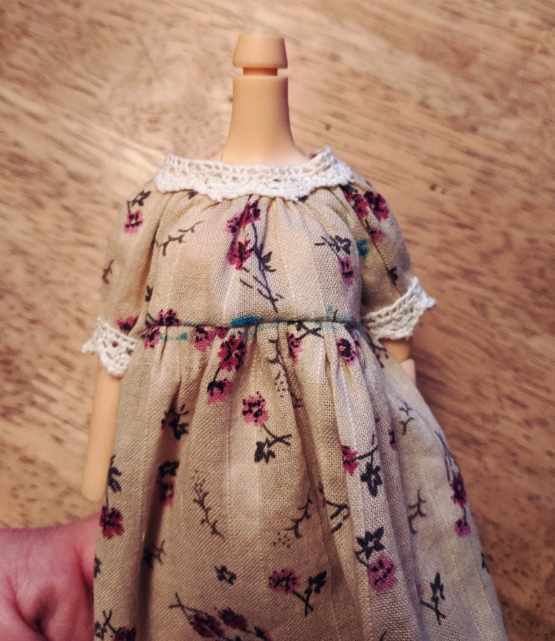 Puede incluir: Un vestido beige con estampado floral y un ribete de encaje blanco alrededor del cuello y las mangas. El vestido tiene una cintura fruncida y una falda suelta y fluida.