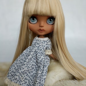 Puede incluir: Una mu&ntilde;eca con el pelo largo y liso rubio y ojos azules que lleva un vestido floral azul y blanco con un borde de encaje.